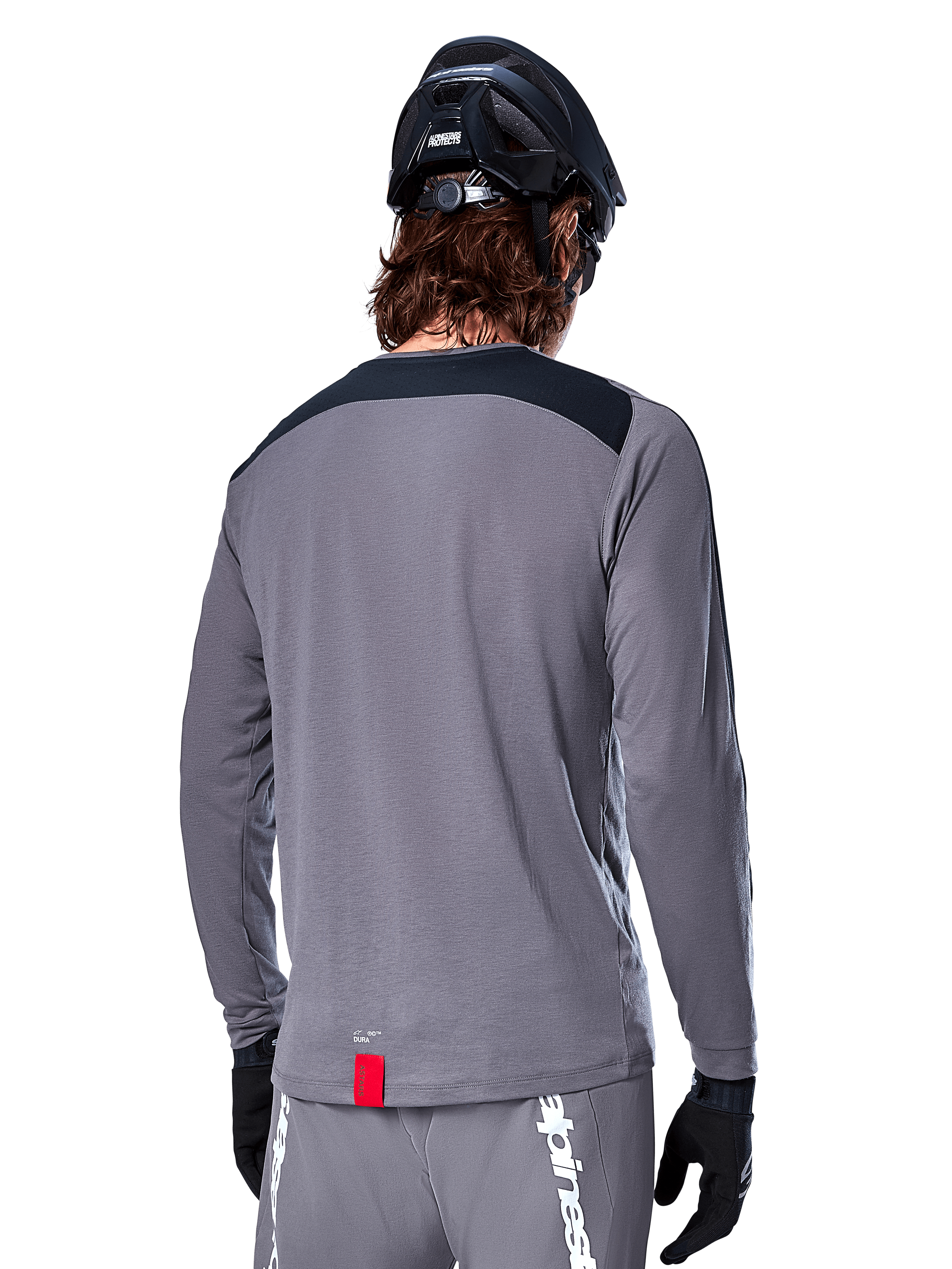 Alpinestars A-Dura Dri Astar Langarmtrikot, dunkelgraues MTB-Trikot von hinten gezeigt, mit schwarzem Schulterbereich und kleinem roten Markenlabel, kombiniert mit schwarzem Mountainbike-Helm und Handschuhen.