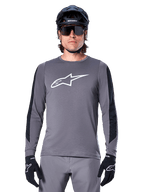 Alpinestars A-Dura Dri Astar Langarmtrikot, Dunkelgrau, mit großem weißen Astar-Logo auf der Brust und schwarzen Mesh-Einsätzen an den Armen, getragen von einem Fahrer mit schwarzem Mountainbike-Helm und Performance-Sonnenbrille, MTB-Radsportbekleidung