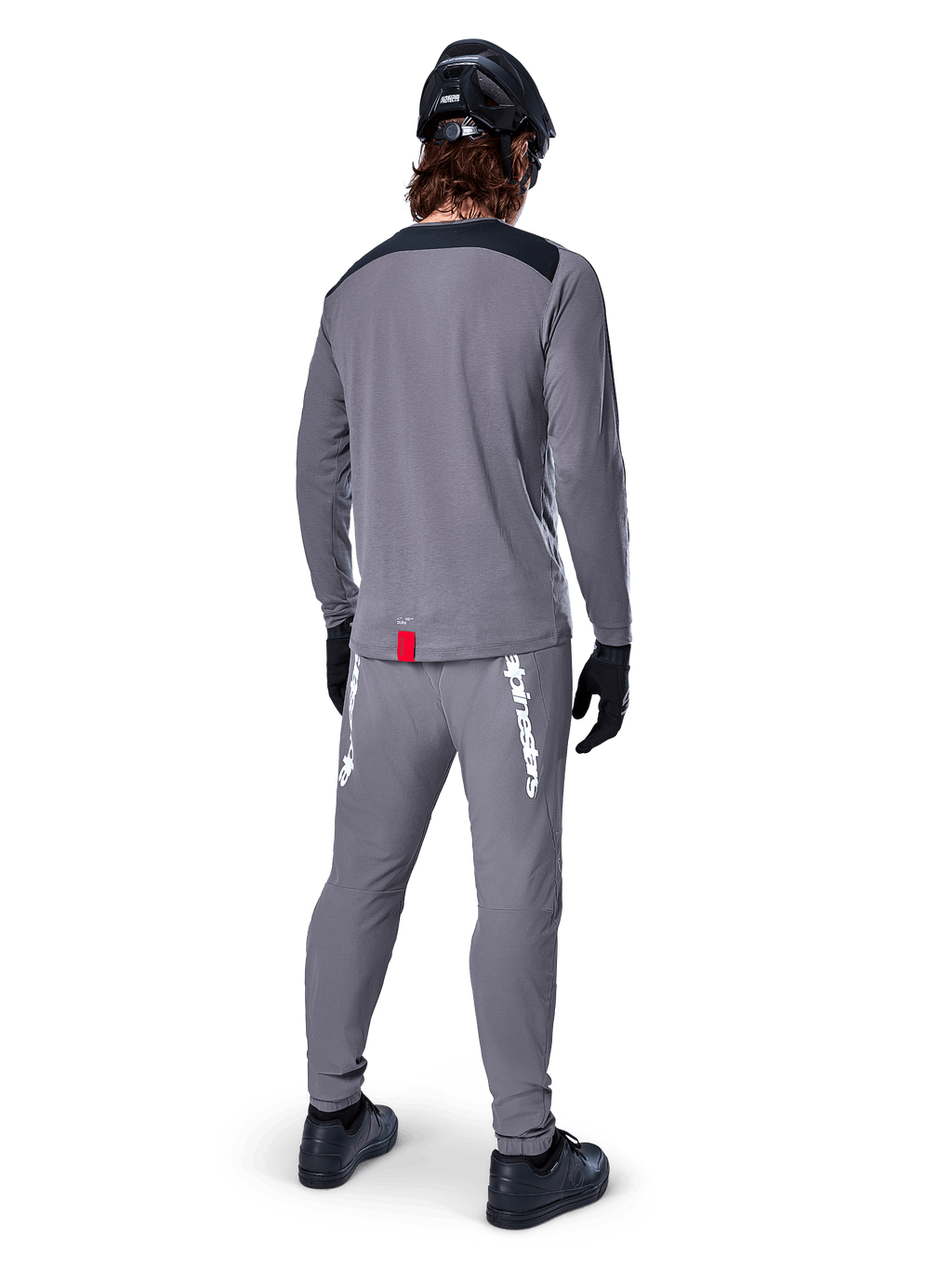 Alpinestars A-Dura Dri Astar Langarmtrikot, Dunkelgrau, Rückansicht eines Fahrers mit passender Hose und schwarzem Helm, mit schwarzen Schultereinsätzen und einem kleinen roten Etikett am Saum, technische Offroad-Radbekleidung