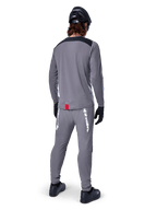 Alpinestars A-Dura Dri Astar Langarmtrikot, Dunkelgrau, Rückansicht eines Fahrers mit passender Hose und schwarzem Helm, mit schwarzen Schultereinsätzen und einem kleinen roten Etikett am Saum, technische Offroad-Radbekleidung