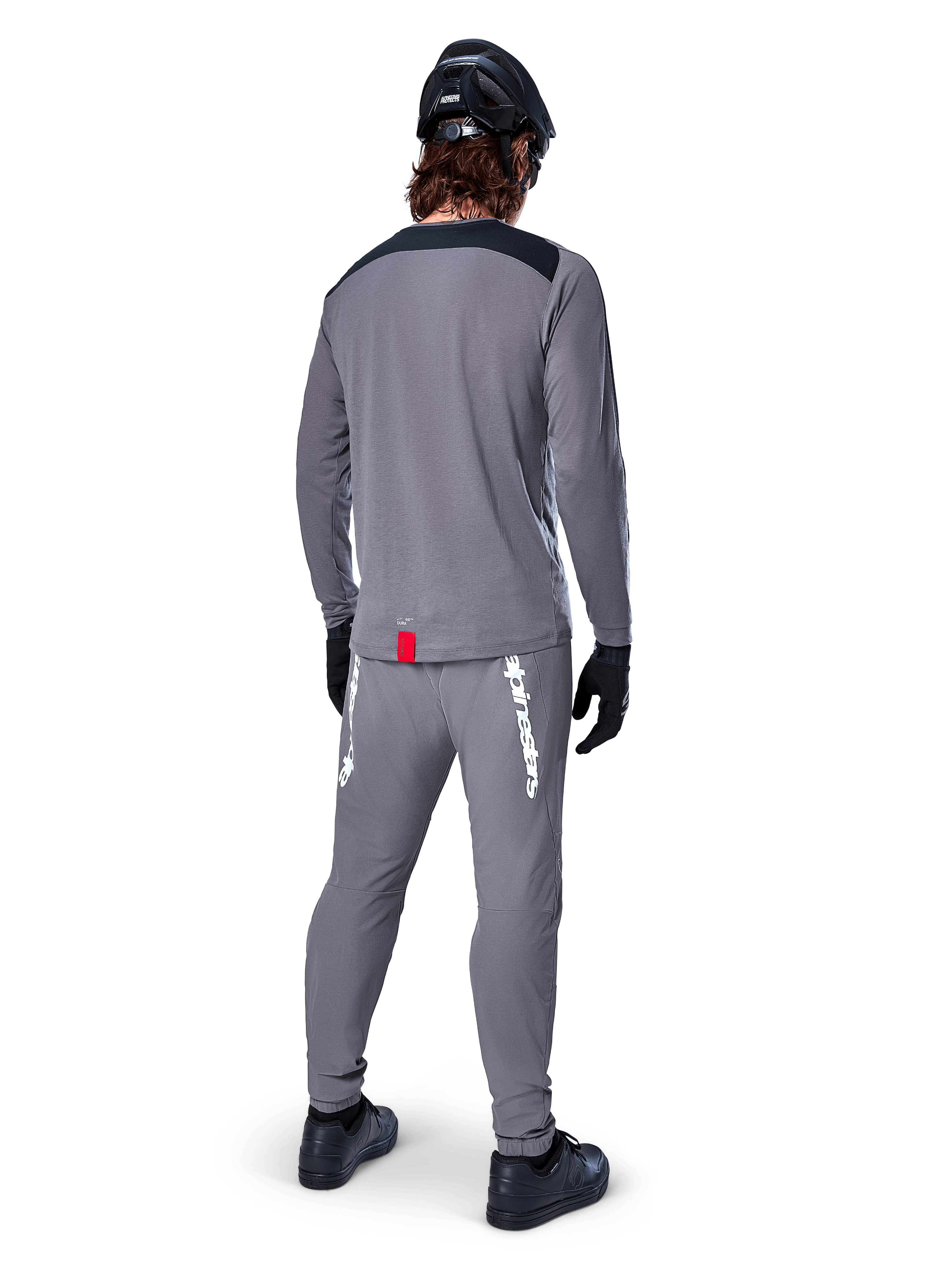 Alpinestars A-Dura Dri Astar Langarmtrikot, Dunkelgrau, Rückansicht eines Fahrers mit passender Hose und schwarzem Helm, mit schwarzen Schultereinsätzen und einem kleinen roten Etikett am Saum, technische Offroad-Radbekleidung