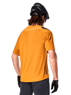 Alpinestars A-Aria Polartec Switch Trikot, Kurzarm-MTB-Trikot, Dunkelgold, Rückansicht mit Performance-Passform, getragen mit schwarzem Helm und grauen Shorts, strapazierfähige und atmungsaktive Trail-Bekleidung