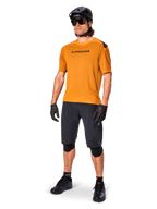 Männlicher Fahrer im Alpinestars A-Aria Polartec Switch Trikot in Dark Gold, kurzärmeliges MTB-Trikot, kombiniert mit schwarzen Radshorts, Knieschonern, schwarzem Helm und Handschuhen, professionelles Mountainbike-Outfit für Trail-Riding