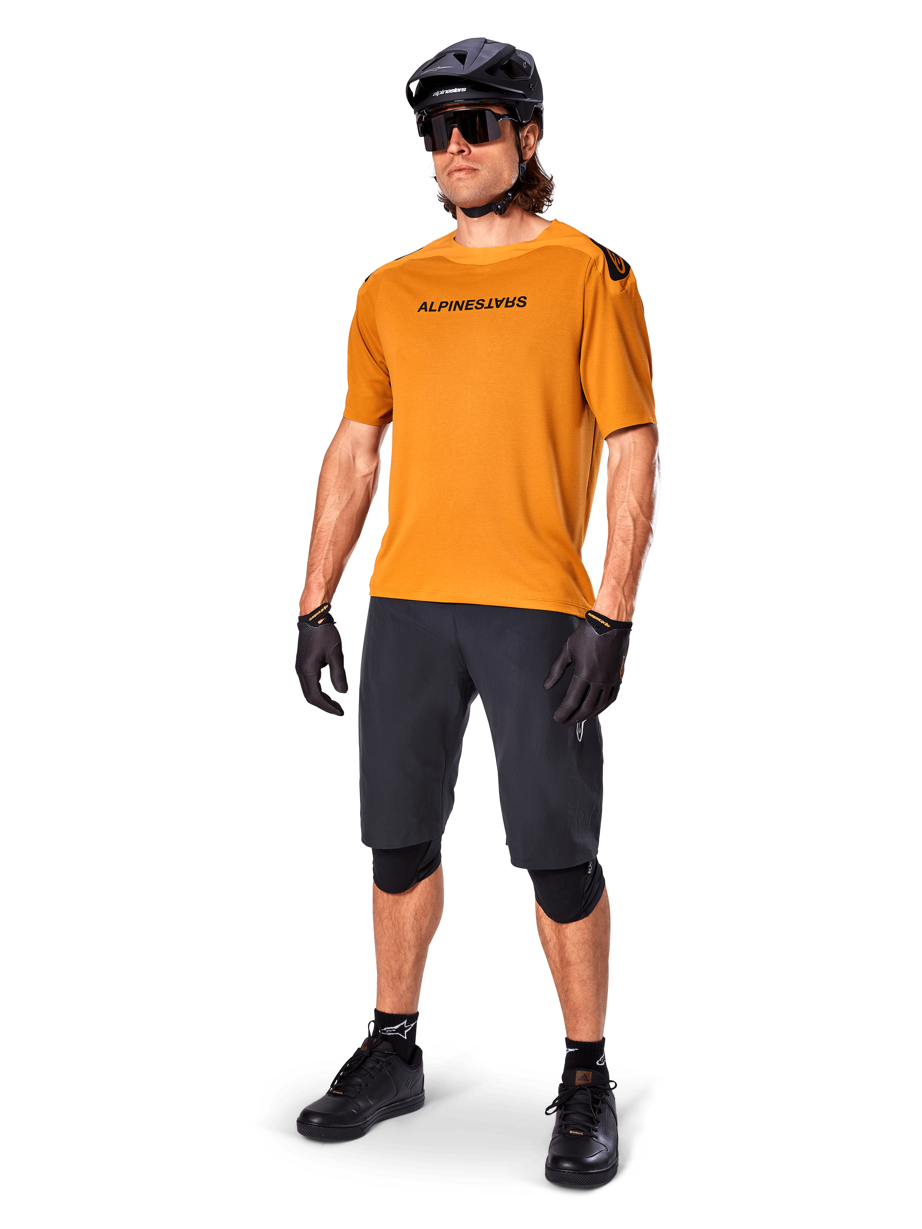 Männlicher Fahrer im Alpinestars A-Aria Polartec Switch Trikot in Dark Gold, kurzärmeliges MTB-Trikot, kombiniert mit schwarzen Radshorts, Knieschonern, schwarzem Helm und Handschuhen, professionelles Mountainbike-Outfit für Trail-Riding