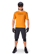 Männlicher Radfahrer im Alpinestars A-Aria Polartec Switch Kurzarmtrikot in Dark Gold, kombiniert mit schwarzen MTB-Shorts, Helm, Sonnenbrille und Handschuhen, professionelle Mountainbike-Trail-Ausrüstung