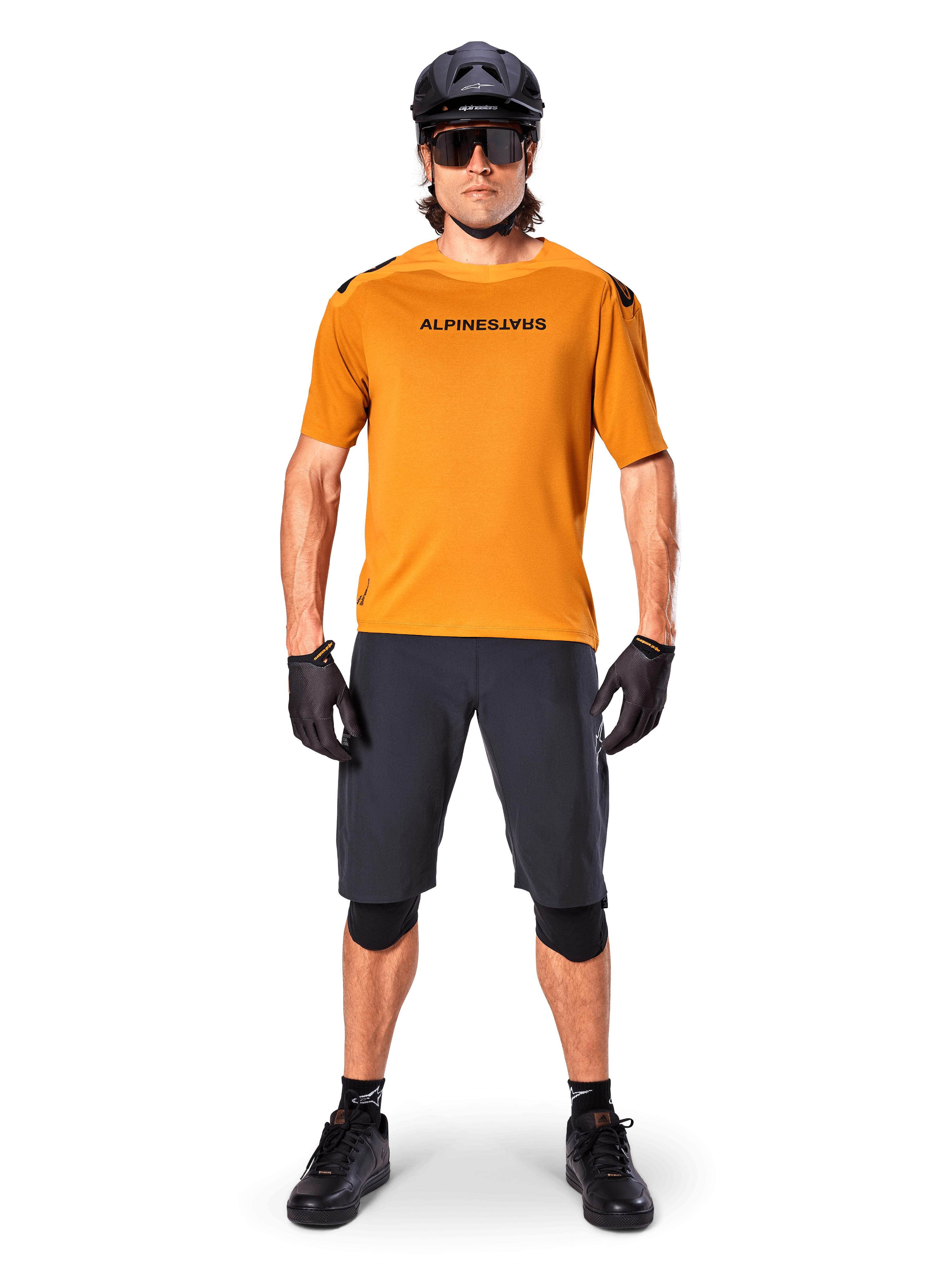 Männlicher Radfahrer im Alpinestars A-Aria Polartec Switch Kurzarmtrikot in Dark Gold, kombiniert mit schwarzen MTB-Shorts, Helm, Sonnenbrille und Handschuhen, professionelle Mountainbike-Trail-Ausrüstung
