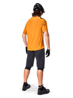 Alpinestars A-Aria Polartec Switch Trikot, kurzärmeliges MTB-Trikot, Dunkelgold, Rückansicht eines Models in dunkelgrauen Mountainbike-Shorts, schwarzem Helm und Handschuhen, technische Radsportbekleidung für Offroad-Trails