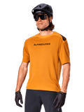 Alpinestars A-Aria Polartec Switch Trikot, Kurzarm-MTB-Trikot, Dunkelgold mit schwarzem Branding, getragen von Model mit schwarzem Helm, Sonnenbrille und Radhandschuhen, Hochleistungs-Mountainbike-Bekleidung
