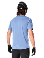 Alpinestars A-Dura Dri Astar Kurzarmtrikot, MTB-Trikot, Infinity Blue, Rückansicht mit Mesh-Belüftungseinsätzen an den Schultern und verlängertem Rücken, kombiniert mit schwarzem MTB-Helm und Handschuhen, Performance-Radbekleidung