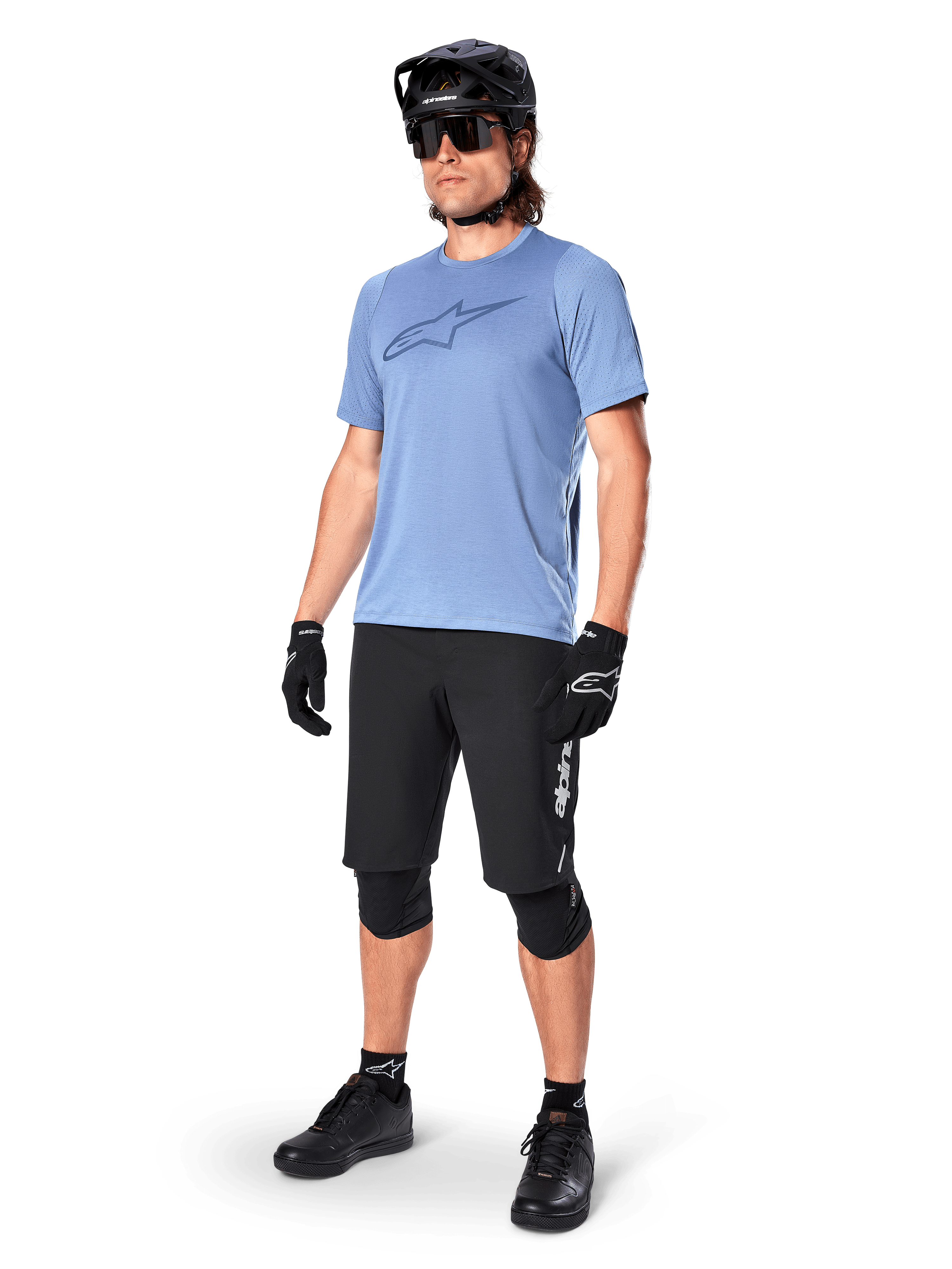 Männlicher Fahrer im Alpinestars A-Dura Dri Astar Kurzarmtrikot in Infinity Blue, kombiniert mit schwarzen MTB-Shorts, schwarzem Helm, Sonnenbrille, Knieschützern und Handschuhen, komplettes Mountainbike-Outfit