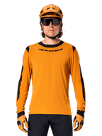 Männliches Model im Alpinestars A-Dura Dri Wool Langarmtrikot in Dark Gold, kombiniert mit einem passenden goldenen MTB-Helm und Handschuhen, schwarze Mesh-Einsätze an Schultern und Ärmeln, schwarzer Alpinestars-Schriftzug auf der Brust, dunkle Sonnenbrille und Hose, technische Mountainbike-Bekleidung