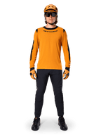 Mountainbiker im Alpinestars A-Dura Dri Wool Langarmtrikot, Dunkelgold mit schwarzen Akzenten an Kragen und Ärmeln, kombiniert mit einer schwarzen Hose, passendem goldenem Helm, Handschuhen und einer schwarzen Sonnenbrille, professionelle Mountainbike-Ausrüstung