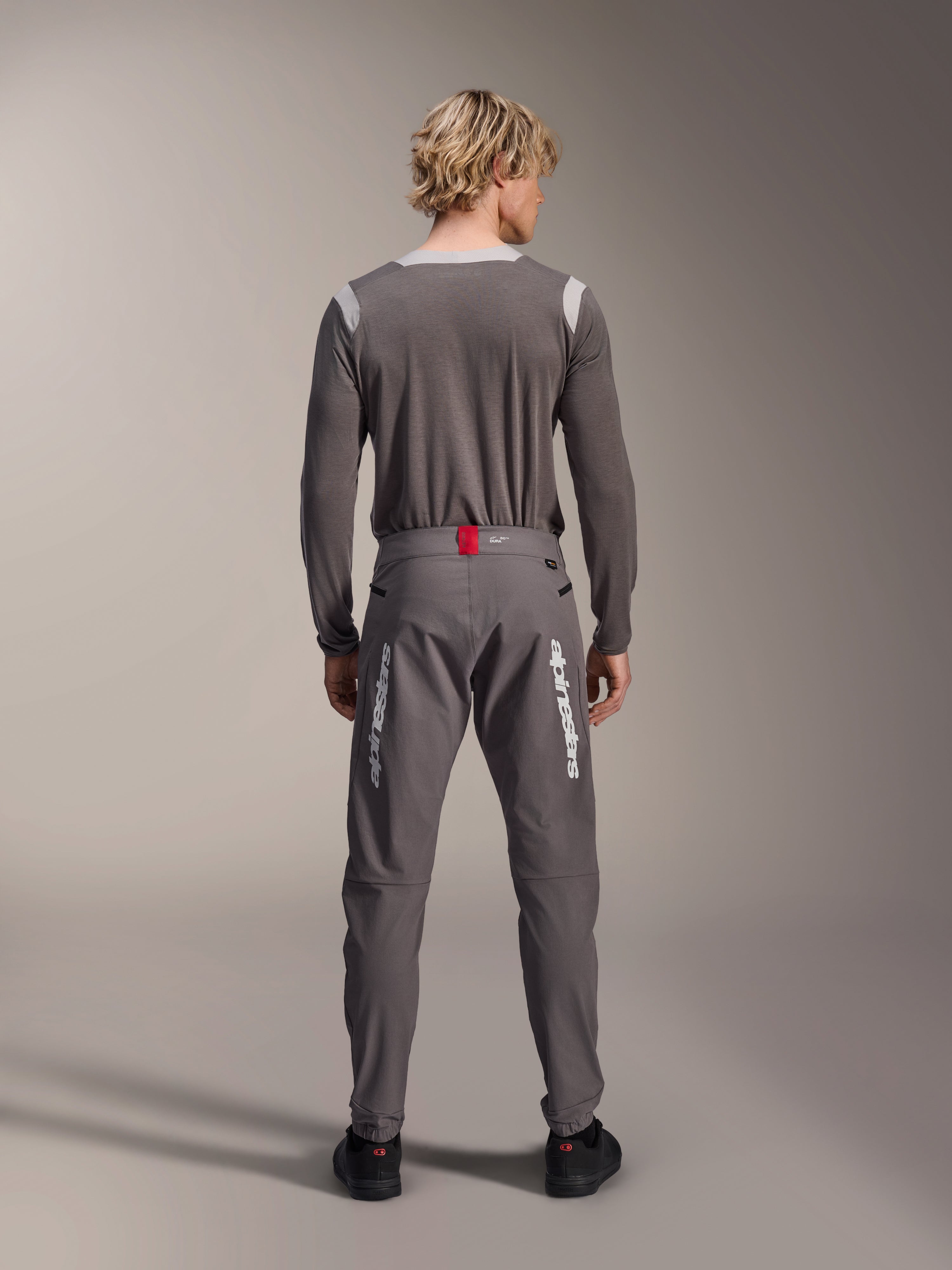 Model in der Rückansicht trägt Alpinestars A-Dura Elite Hose in Dunkelgrau und ein passendes graues Langarm-Trikot, weiße Alpinestars-Logos auf den Beinen, roter Akzent am Bund, schwarze Schuhe, professionelle MTB-Ausrüstung.