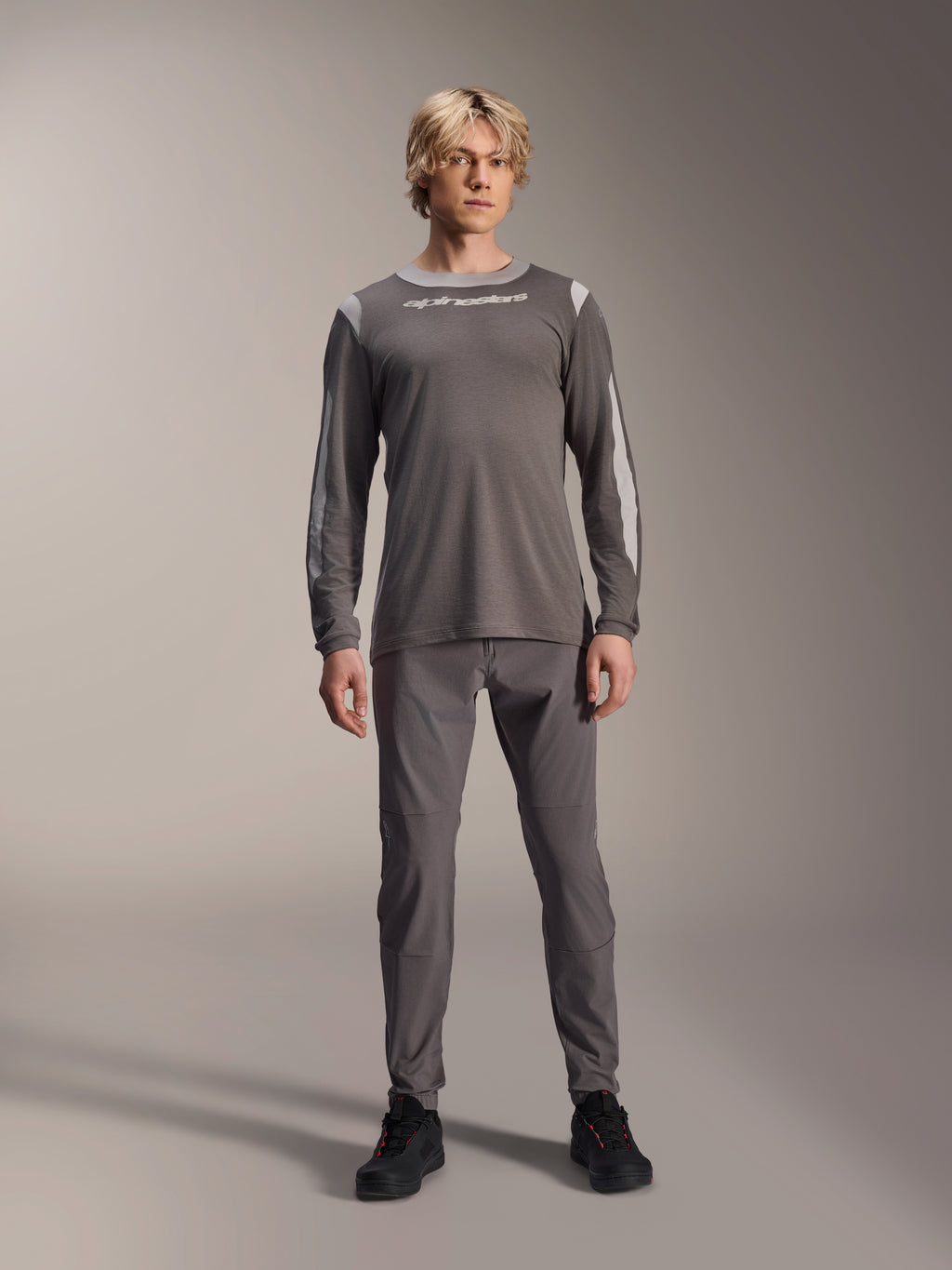 Alpinestars A-Dura Dri Wool Langarmtrikot, Dunkelgrau, getragen von einem Modell mit passender grauer Hose und schwarzen Schuhen, weißes Alpinestars-Branding auf der Brust, hellgraue Mesh-Einsätze an Schultern und Ärmeln, technische Mountainbike-Ausrüstung