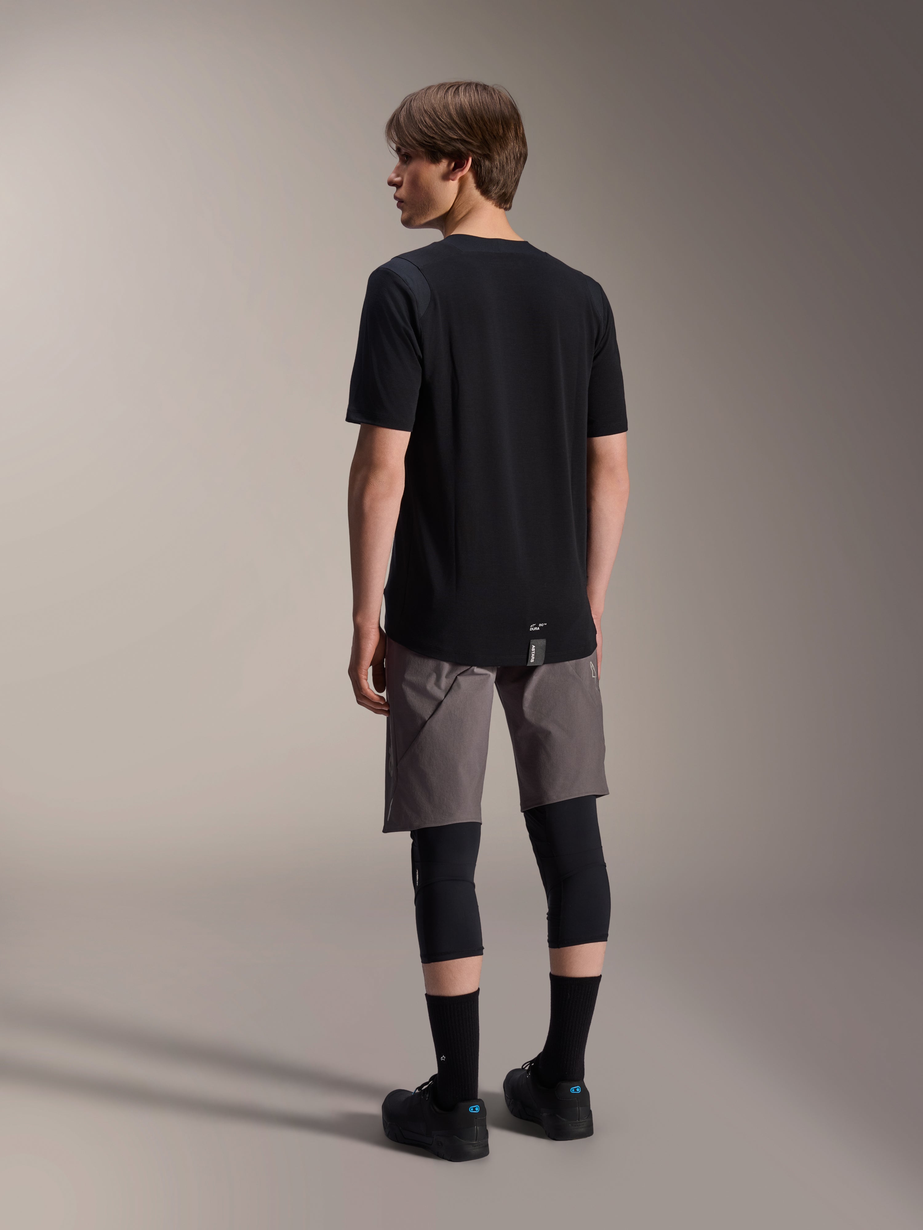 Alpinestars A-Dura Dri Wool Kurzarmtrikot, Schwarz, Rückansicht am Modell, getragen mit grauen Mountainbike-Shorts und schwarzen Knieprotektoren, mit verlängertem Rückensaum und dezentem Logo, technische MTB-Bekleidung