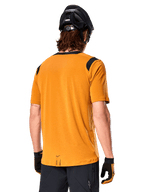 Alpinestars A-Dura Dri Wool Kurzarmtrikot, Dunkelgold, Rückansicht eines Mannes, der das Trikot mit schwarzen Mesh-Einsätzen an den Schultern trägt, kombiniert mit schwarzem MTB-Helm und Handschuhen, MTB-Ausrüstung in regulärer Passform