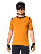 Alpinestars A-Dura Dri Wool Kurzarmtrikot, Dark Gold, getragen mit schwarzem MTB-Helm, Sonnenbrille und Handschuhen, mit schwarzem Kragen und Schultereinsätzen, atmungsaktives drirelease-Wollmischgewebe für Mountainbiking