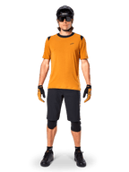 Alpinestars A-Dura Dri Wool Kurzarmtrikot in Dark Gold, Mountainbike-Trikot kombiniert mit schwarzen Shorts und Schutzausrüstung, senfgelbes Material mit schwarzem Kragen und Schulterakzenten, Performance-Radbekleidung für Trail-Fahrten.