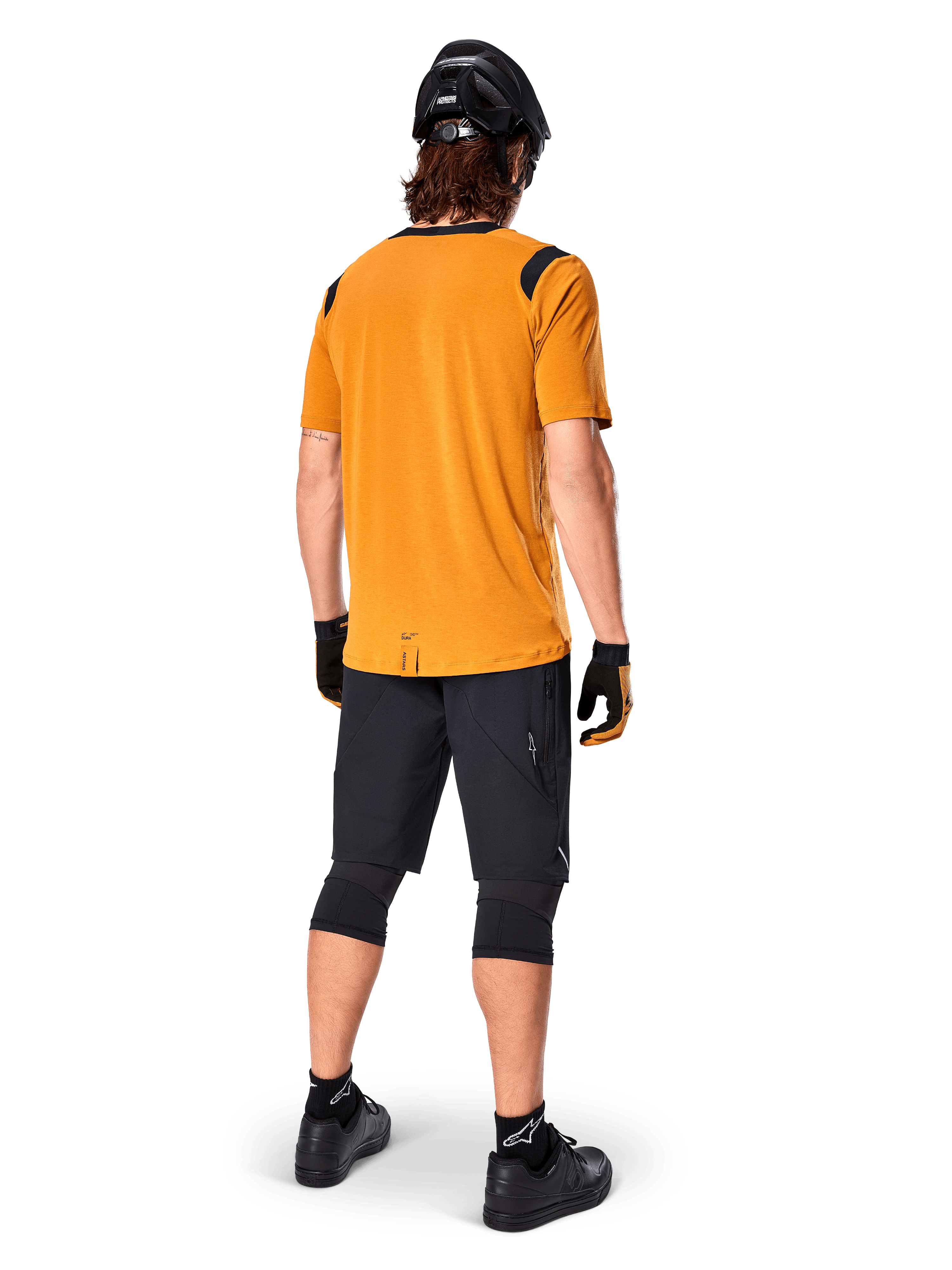 Alpinestars A-Dura Dri Wool Kurzarmtrikot, Radsport-Trikot, Dunkelgold, Rückansicht eines Mountainbikers in senfgelbem Trikot mit schwarzen Mesh-Schulterakzenten, kombiniert mit schwarzen Shorts, Knieschützern, schwarzem Helm und Handschuhen, technische Offroad-Radsportausrüstung