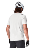 Alpinestars A-Aria Astar Trikot, Kurzarm, White Dove, Rückansicht eines leichten, atmungsaktiven Mountainbike-Trikots, getragen mit einem weißen Helm und schwarzen Fahrradhandschuhen, mit verlängertem Saum für zusätzliche Abdeckung