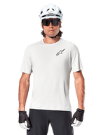 Radfahrer trägt Alpinestars A-Aria Astar Kurzarmtrikot, White Dove mit kleinem schwarzem Astar-Logo auf der Brust, weißer Fahrradhelm, schwarze Sonnenbrille und schwarz-weiße Handschuhe, Performance-MTB-Ausrüstung für Offroad-Fahrten