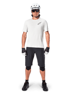 Männlicher Radfahrer in Alpinestars A-Aria Astar Kurzarmtrikot in White Dove, mit schwarzem Astars-Logo auf der Brust, kombiniert mit schwarzen Mountainbike-Shorts, weißem Helm, Sonnenbrille, schwarzen Knieprotektoren und Handschuhen, komplettes Offroad-Radsport-Outfit