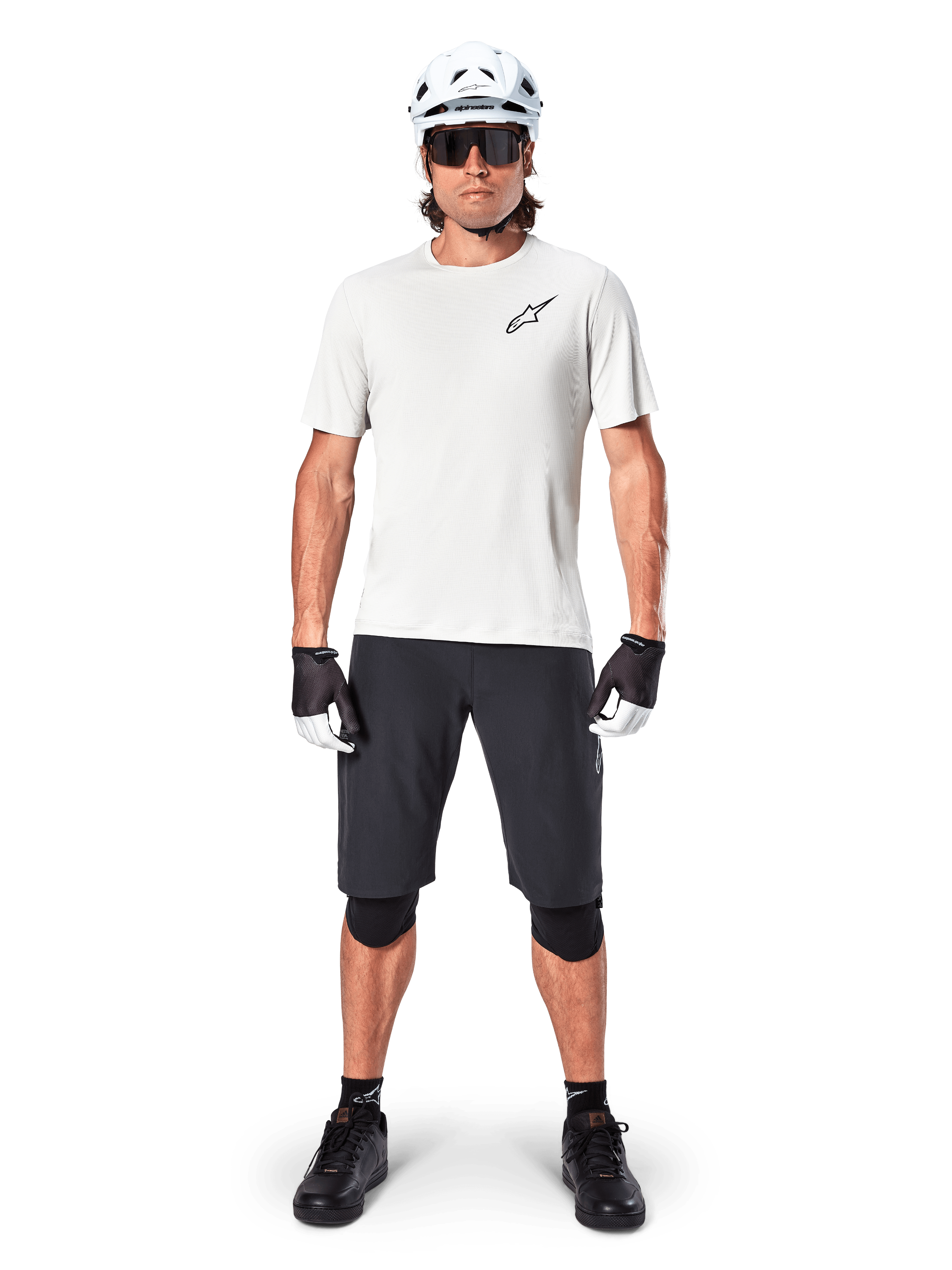 Männlicher Radfahrer in Alpinestars A-Aria Astar Kurzarmtrikot in White Dove, mit schwarzem Astars-Logo auf der Brust, kombiniert mit schwarzen Mountainbike-Shorts, weißem Helm, Sonnenbrille, schwarzen Knieprotektoren und Handschuhen, komplettes Offroad-Radsport-Outfit