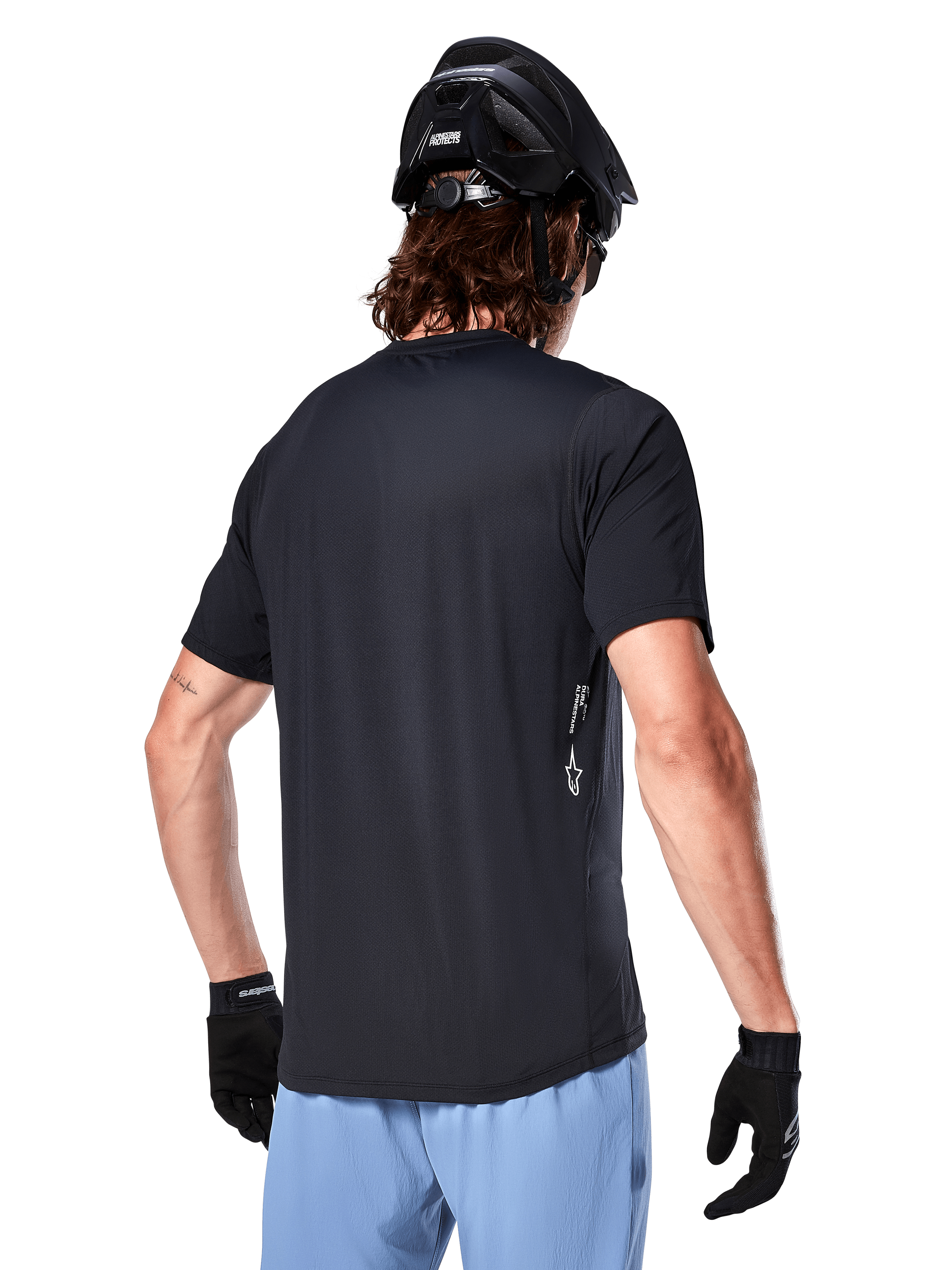 Alpinestars A-Dura Oscar Trikot, Kurzarm-Fahrradtrikot, Schwarz, Rückansicht am Fahrer, kombiniert mit hellblauen Shorts und schwarzem MTB-Helm, kleines weißes Logo an der Seite, Performance-Bekleidung für Mountainbiking