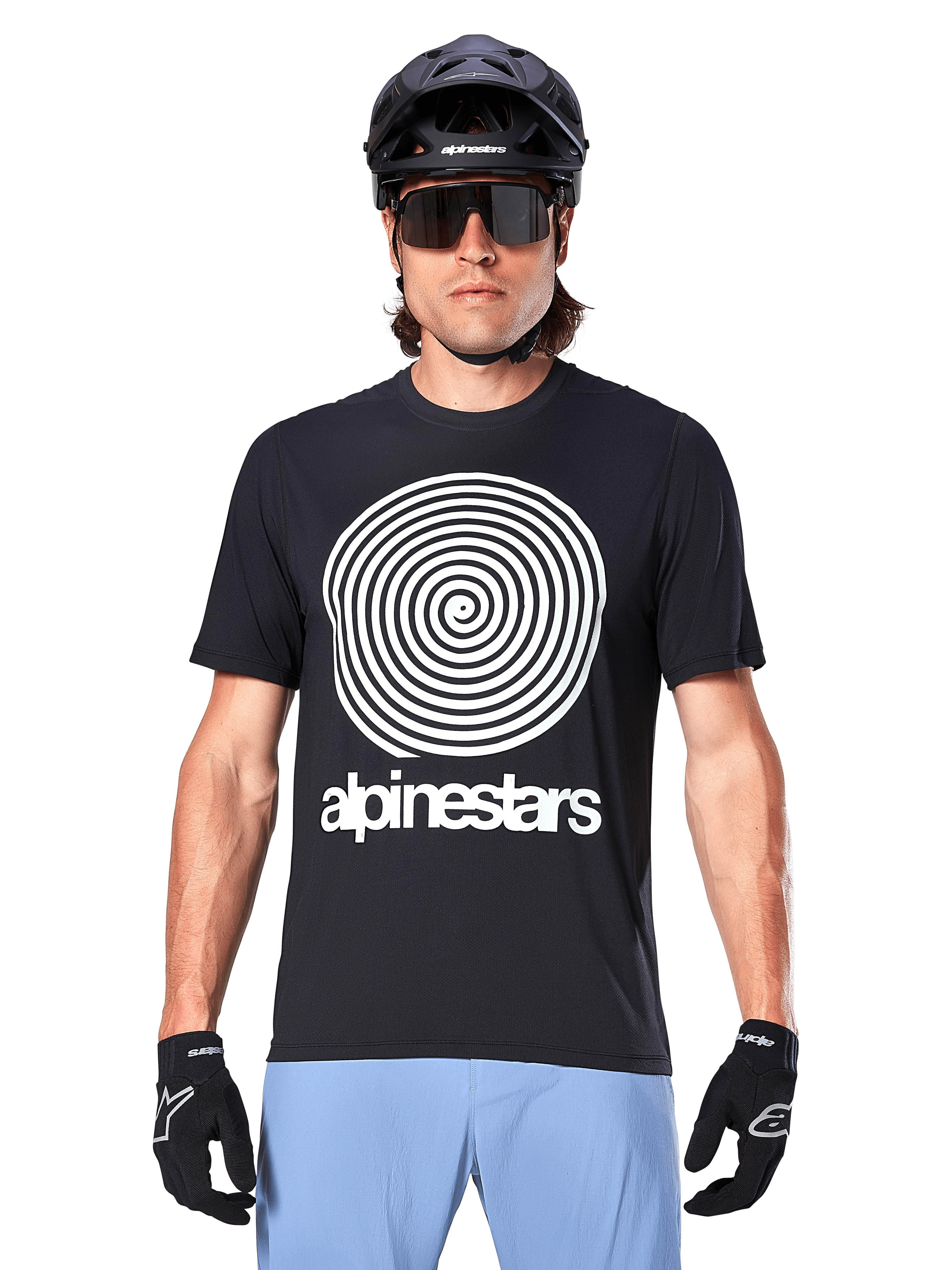 Alpinestars A-Dura Oscar Trikot, Kurzarm-MTB-Trikot, Schwarz, mit einer großen weißen Vintage-Oscar-Spiralgrafik auf der Brust, getragen mit einem schwarzen Fahrradhelm, Sonnenbrille, schwarzen Handschuhen und hellblauen Shorts, vom Heritage-Stil inspirierte Offroad-Fahrradausrüstung
