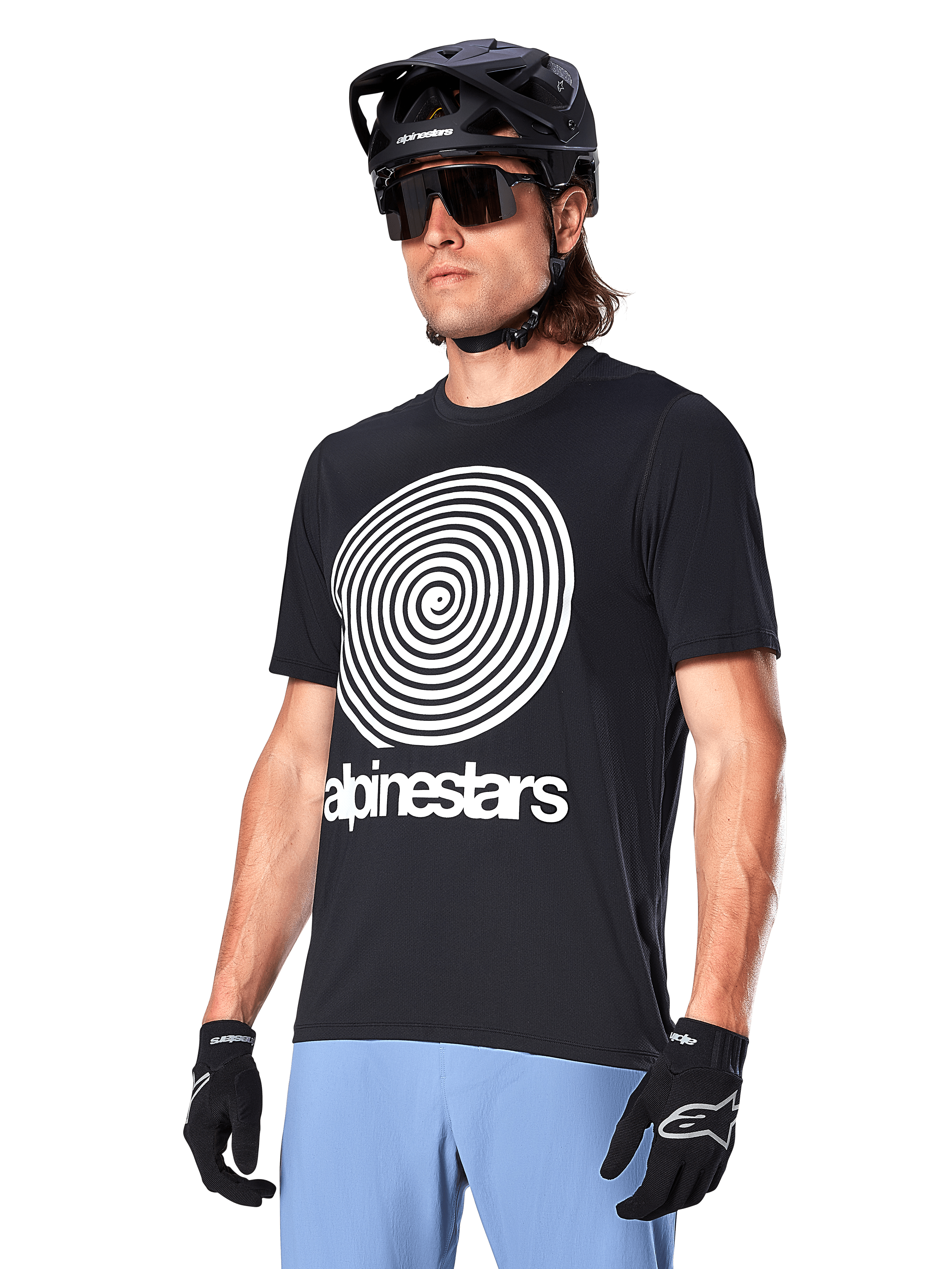 Alpinestars A-Dura Oscar Kurzarmtrikot, Schwarz, mit großer weißer Spiralgrafik und Alpinestars-Schriftzug auf der Brust, getragen von einem Fahrer mit schwarzem MTB-Helm, Sonnenbrille und hellblauer Hose