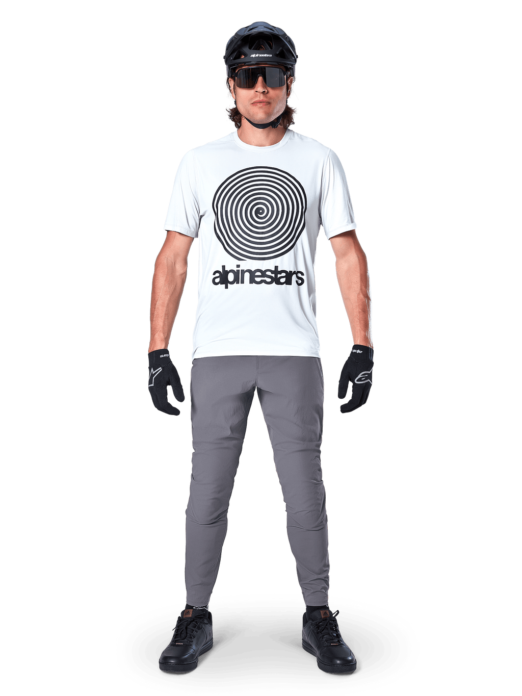 Mountainbiker im Alpinestars A-Dura Oscar Kurzarmtrikot, White Dove mit großem schwarzem Spiral-Logo, kombiniert mit grauer Hose, schwarzem Helm, Sonnenbrille und schwarzen Handschuhen, Performance-MTB-Trail-Ausrüstung