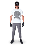 Mountainbiker im Alpinestars A-Dura Oscar Kurzarmtrikot, White Dove mit großem schwarzem Spiral-Logo, kombiniert mit grauer Hose, schwarzem Helm, Sonnenbrille und schwarzen Handschuhen, Performance-MTB-Trail-Ausrüstung