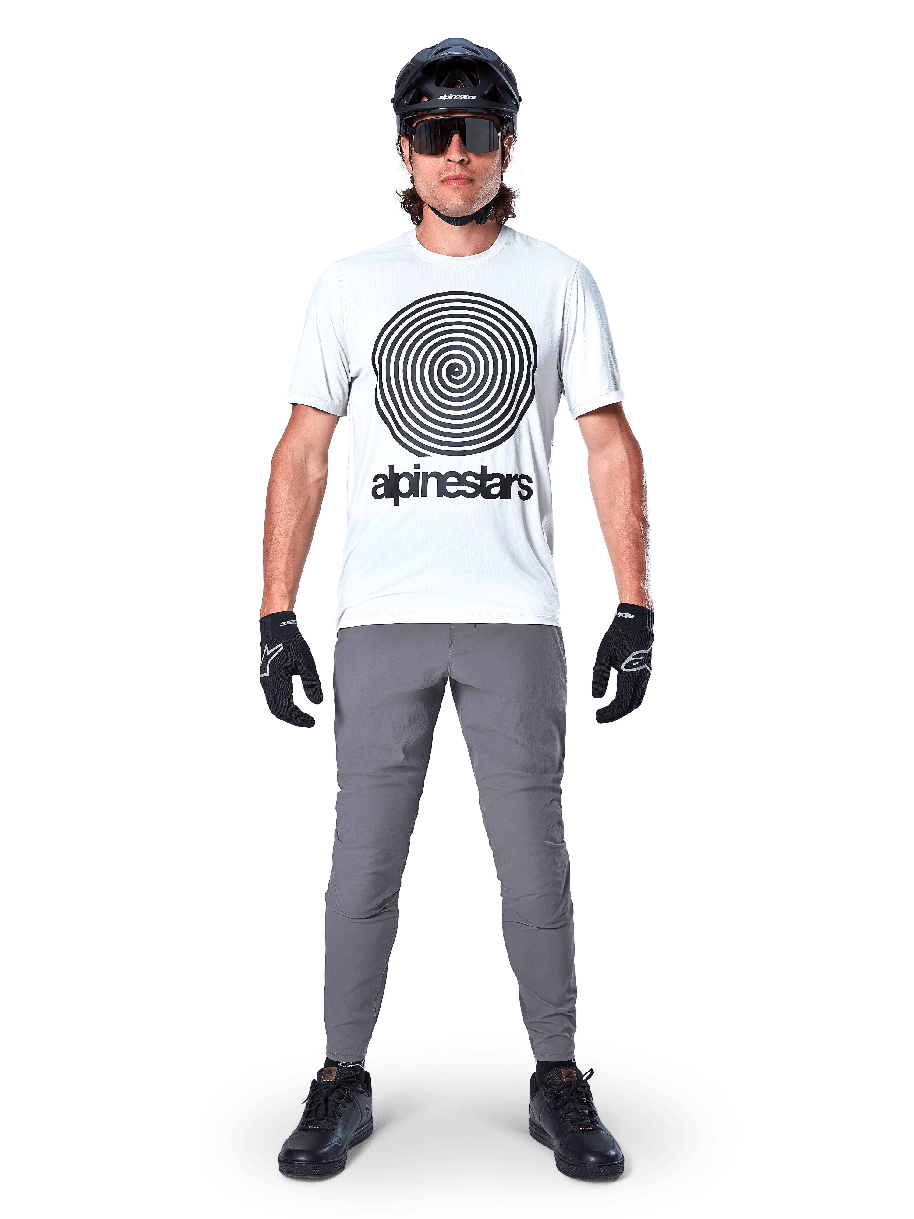 Mountainbiker im Alpinestars A-Dura Oscar Kurzarmtrikot, White Dove mit großem schwarzem Spiral-Logo, kombiniert mit grauer Hose, schwarzem Helm, Sonnenbrille und schwarzen Handschuhen, Performance-MTB-Trail-Ausrüstung