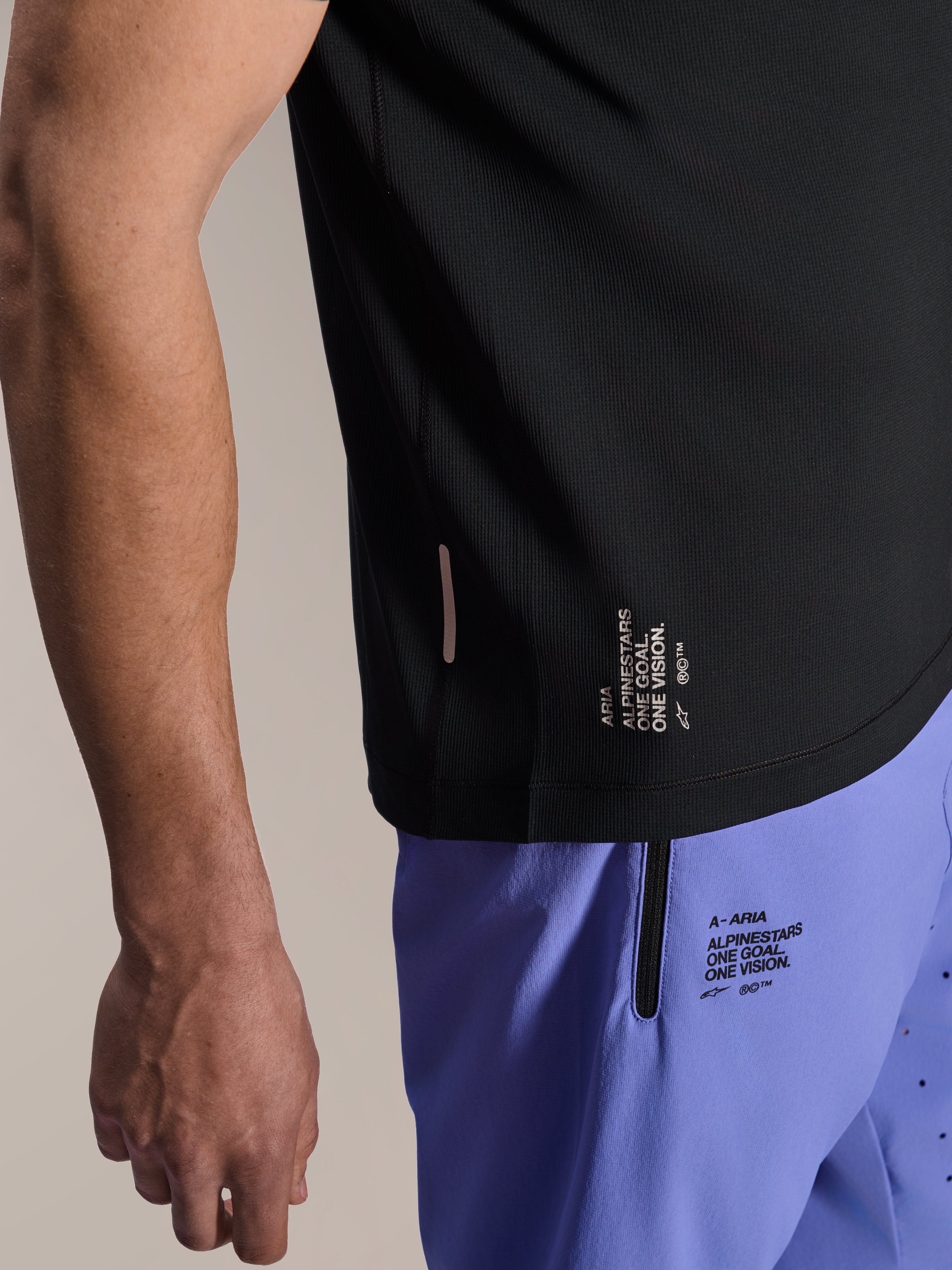 Alpinestars A-Aria Switch Kurzarmtrikot, schwarzes MTB-Trikot, Nahaufnahme des seitlichen Saums mit weißem „ONE GOAL. ONE VISION.“ Branding, getragen mit helllilanen Radshorts mit passenden Logos