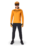 Alpinestars A-Dura Rocker Langarmtrikot in Dark Gold, mit schwarzem Siebdruck-Logo auf der Brust, getragen mit einem passenden goldfarbenen MTB-Helm, schwarzer Sonnenbrille und schwarzer Hose, professionelle Mountainbike-Ausrüstung