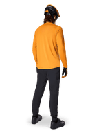 Mountainbike-Fahrer von hinten gesehen im Alpinestars A-Dura Rocker Langarmtrikot in Dark Gold, schwarzer Hose, orange-schwarzem Helm und schwarzen Schuhen, koordinierte Performance-MTB-Ausrüstung für Trailfahrten