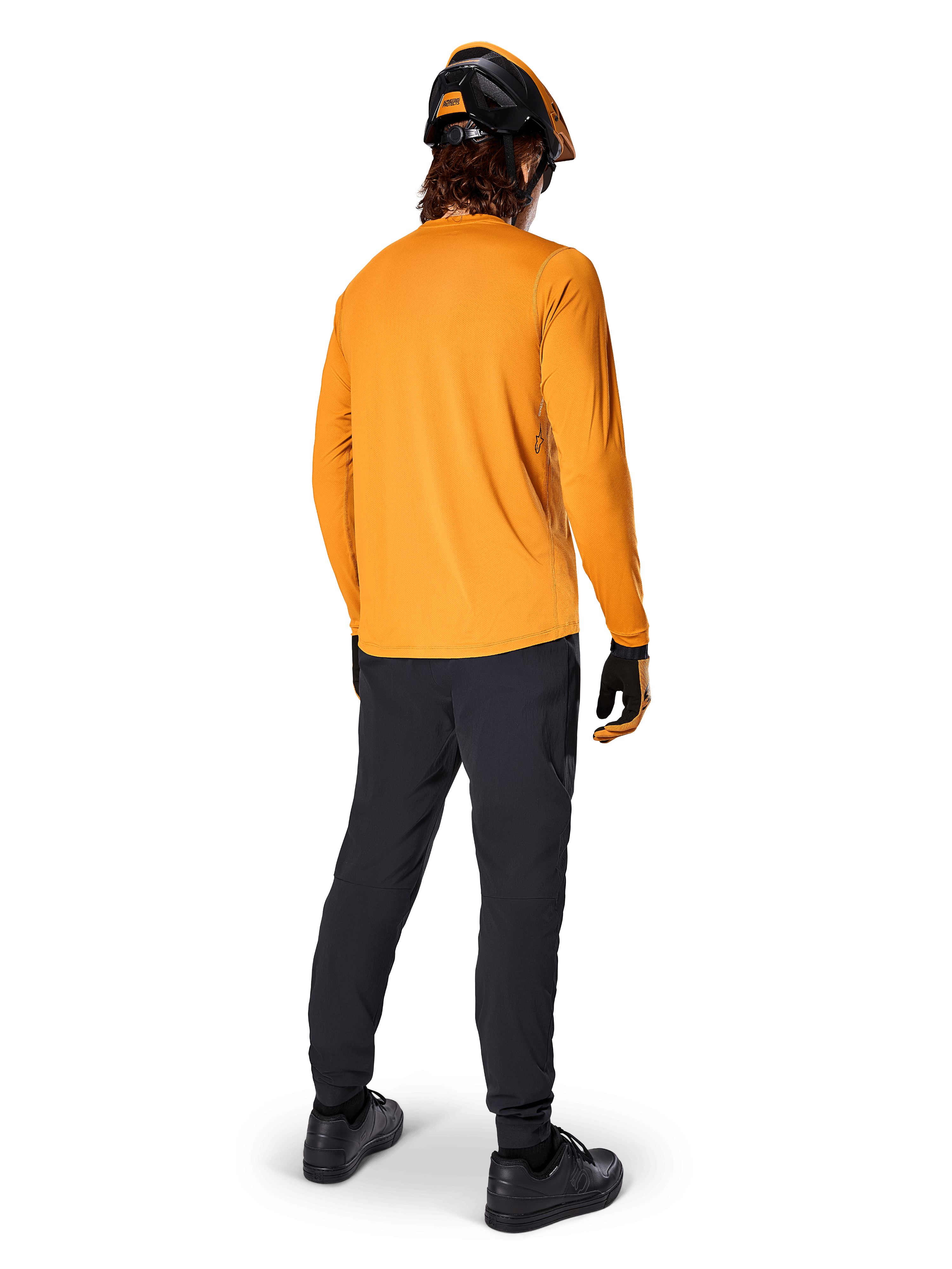 Mountainbike-Fahrer von hinten gesehen im Alpinestars A-Dura Rocker Langarmtrikot in Dark Gold, schwarzer Hose, orange-schwarzem Helm und schwarzen Schuhen, koordinierte Performance-MTB-Ausrüstung für Trailfahrten