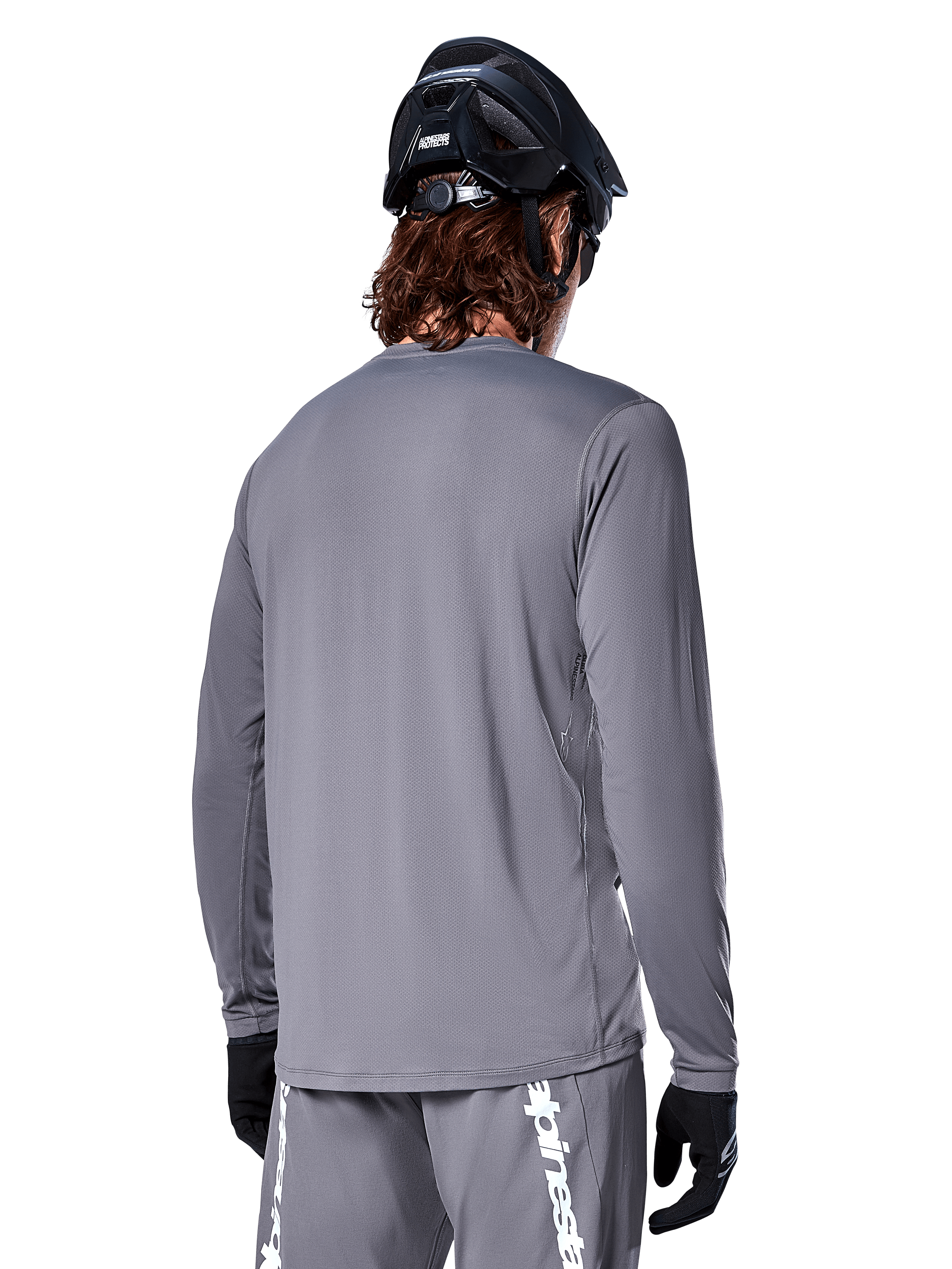 Alpinestars A-Dura Rocker Langarmtrikot in Dunkelgrau, Rückansicht eines Mountainbikers, kombiniert mit passenden grauen Shorts mit weißem Branding, schwarzem MTB-Helm und schwarzen Handschuhen, professionelle Offroad-Radsportausrüstung.