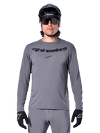 Männlicher Fahrer im Alpinestars A-Dura Rocker Langarmtrikot, dunkelgraues MTB-Trikot mit schwarzem Schriftzug auf der Brust, kombiniert mit grauer Hose, schwarzem Helm und Handschuhen, konzipiert für den Offroad-Radsport