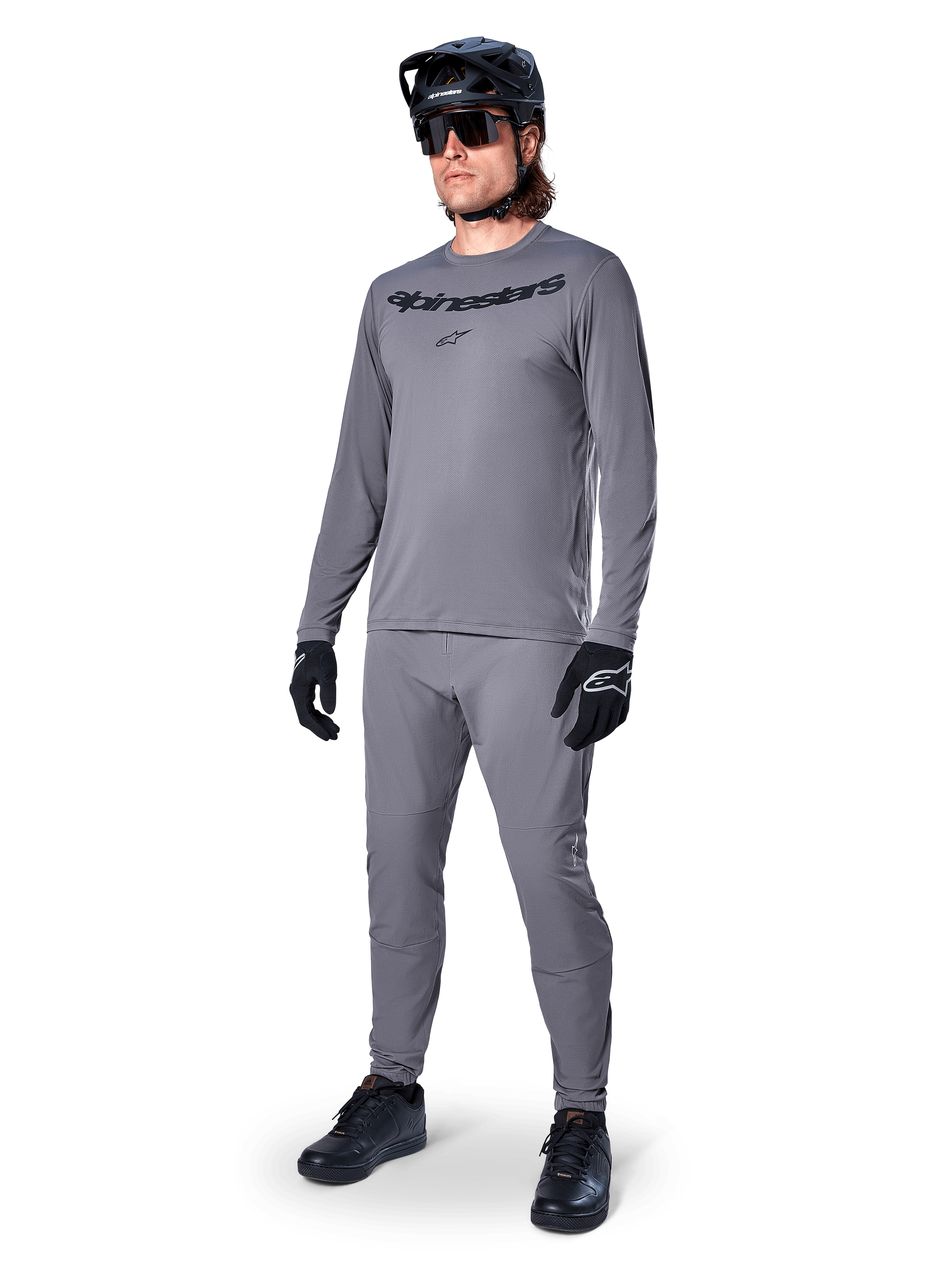 Alpinestars A-Dura Rocker Langarmtrikot, Dunkelgrau, getragen von einem Modell mit passender grauer Hose, schwarzem Mountainbike-Helm, Sonnenbrille und Handschuhen, lässiger MTB-Stil für Geländefahrten
