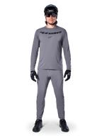 Männlicher Radfahrer im Alpinestars A-Dura Rocker Langarmtrikot in Dunkelgrau, passende graue Hose, schwarzer MTB-Helm, Sonnenbrille und Handschuhe, professionelle Mountainbike-Bekleidung für Offroad-Trails