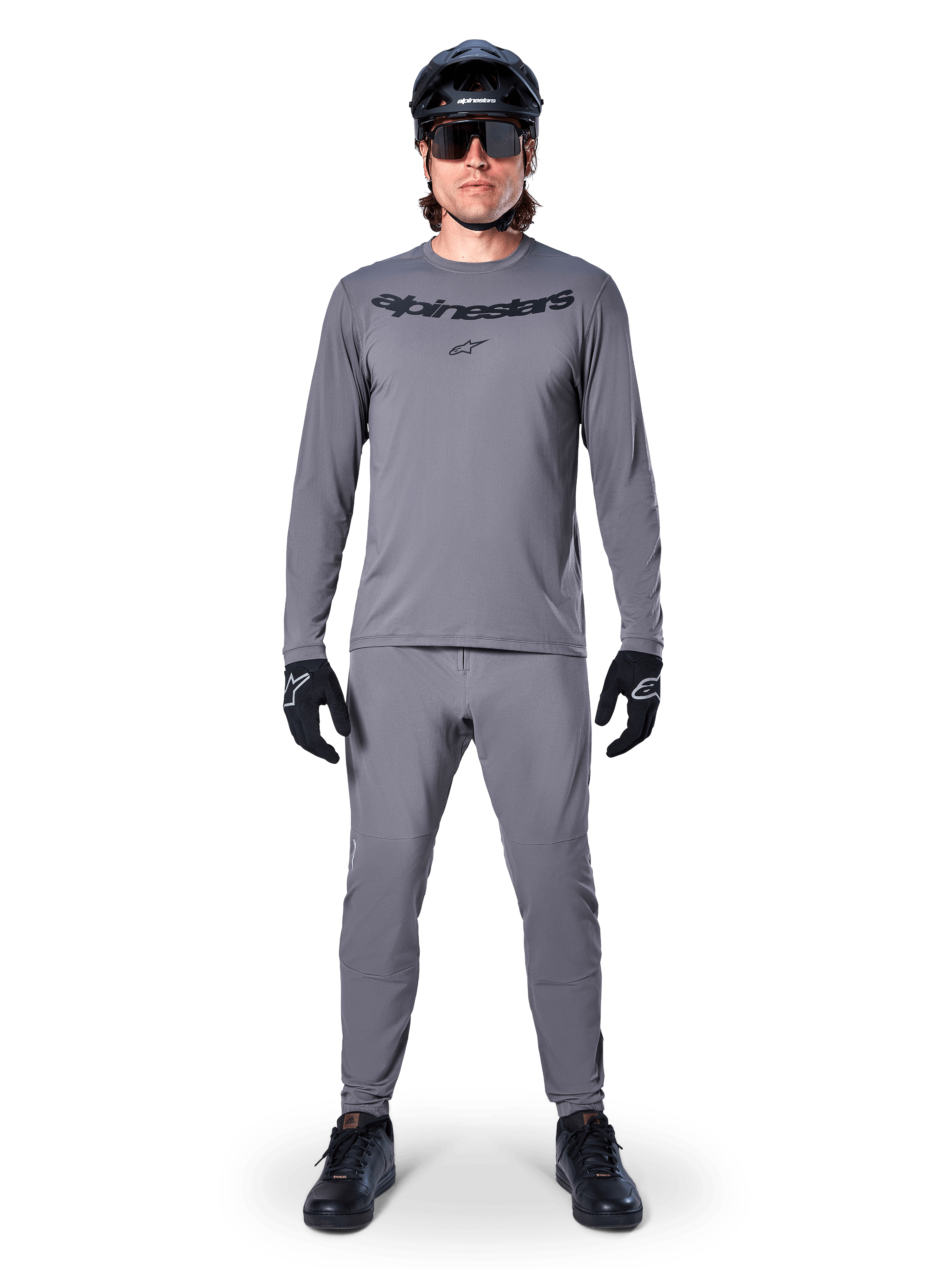 Männlicher Radfahrer im Alpinestars A-Dura Rocker Langarmtrikot in Dunkelgrau, passende graue Hose, schwarzer MTB-Helm, Sonnenbrille und Handschuhe, professionelle Mountainbike-Bekleidung für Offroad-Trails