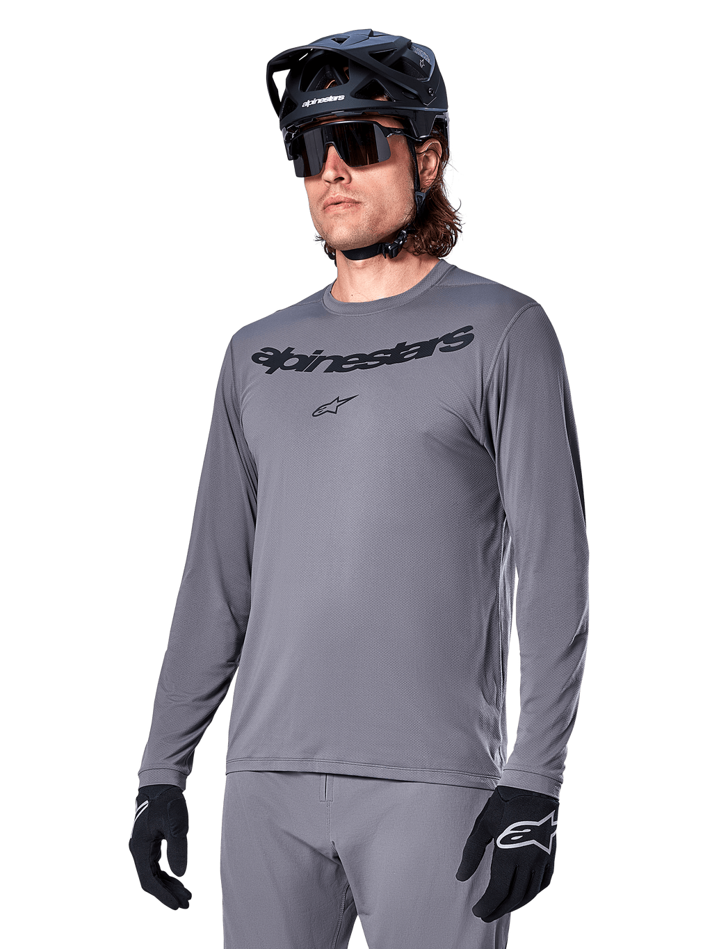 Alpinestars A-Dura Rocker Langarmtrikot, graues MTB-Mountainbike-Trikot, getragen mit schwarzem Helm und Handschuhen, mit schwarzem Schriftzug auf der Brust, sportliche Radbekleidung für Offroad-Fahrten, schlichter Hintergrund