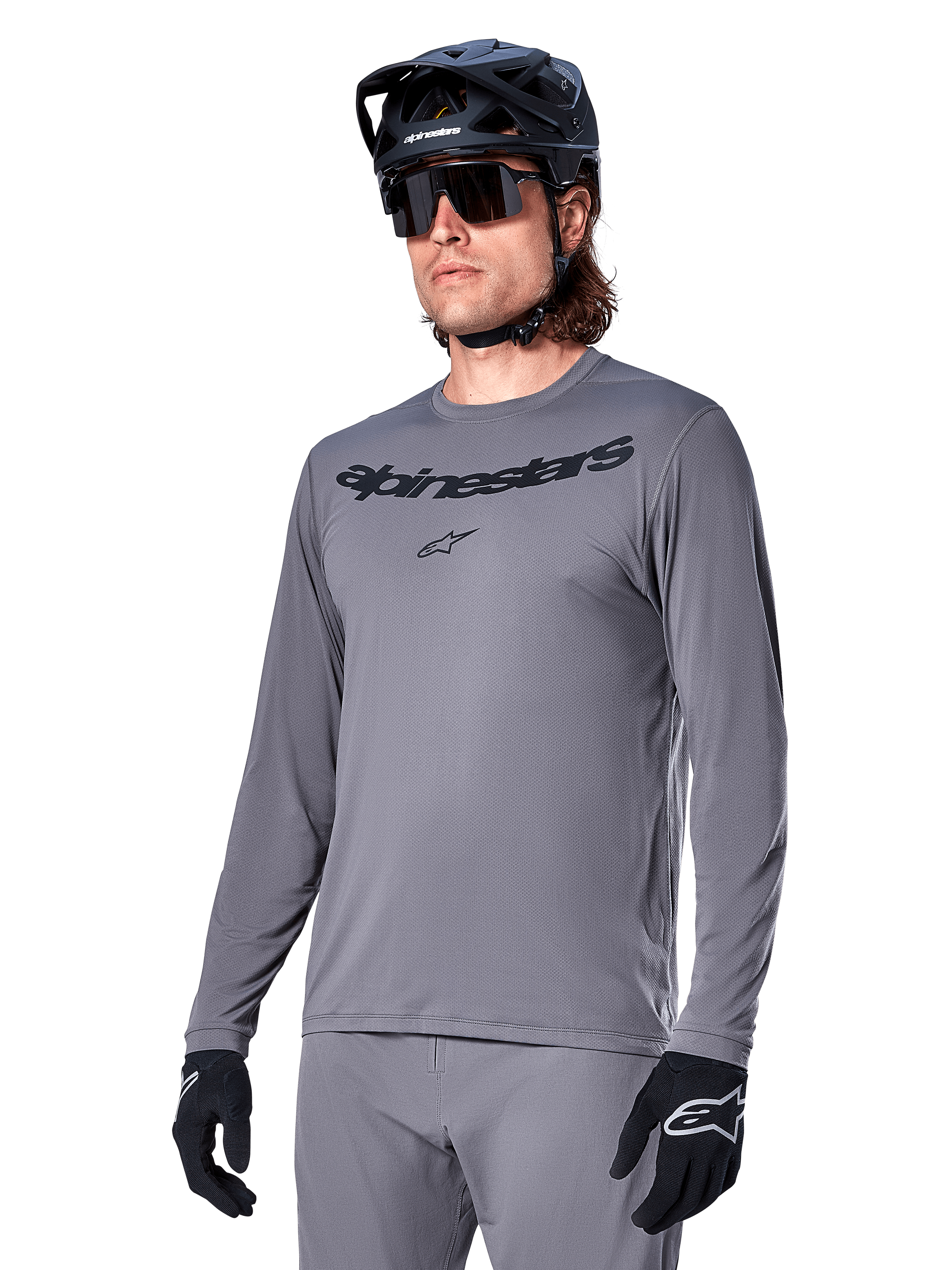 Alpinestars A-Dura Rocker Langarmtrikot, graues MTB-Mountainbike-Trikot, getragen mit schwarzem Helm und Handschuhen, mit schwarzem Schriftzug auf der Brust, sportliche Radbekleidung für Offroad-Fahrten, schlichter Hintergrund