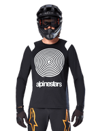 Alpinestars A-Supra Oscar Long Sleeve Jersey, Black with white spiral graphic and shoulder accents, worn by a rider with a black full-face helmet and goggles, black and orange pants, performance cycling gear for MTB gravity riding