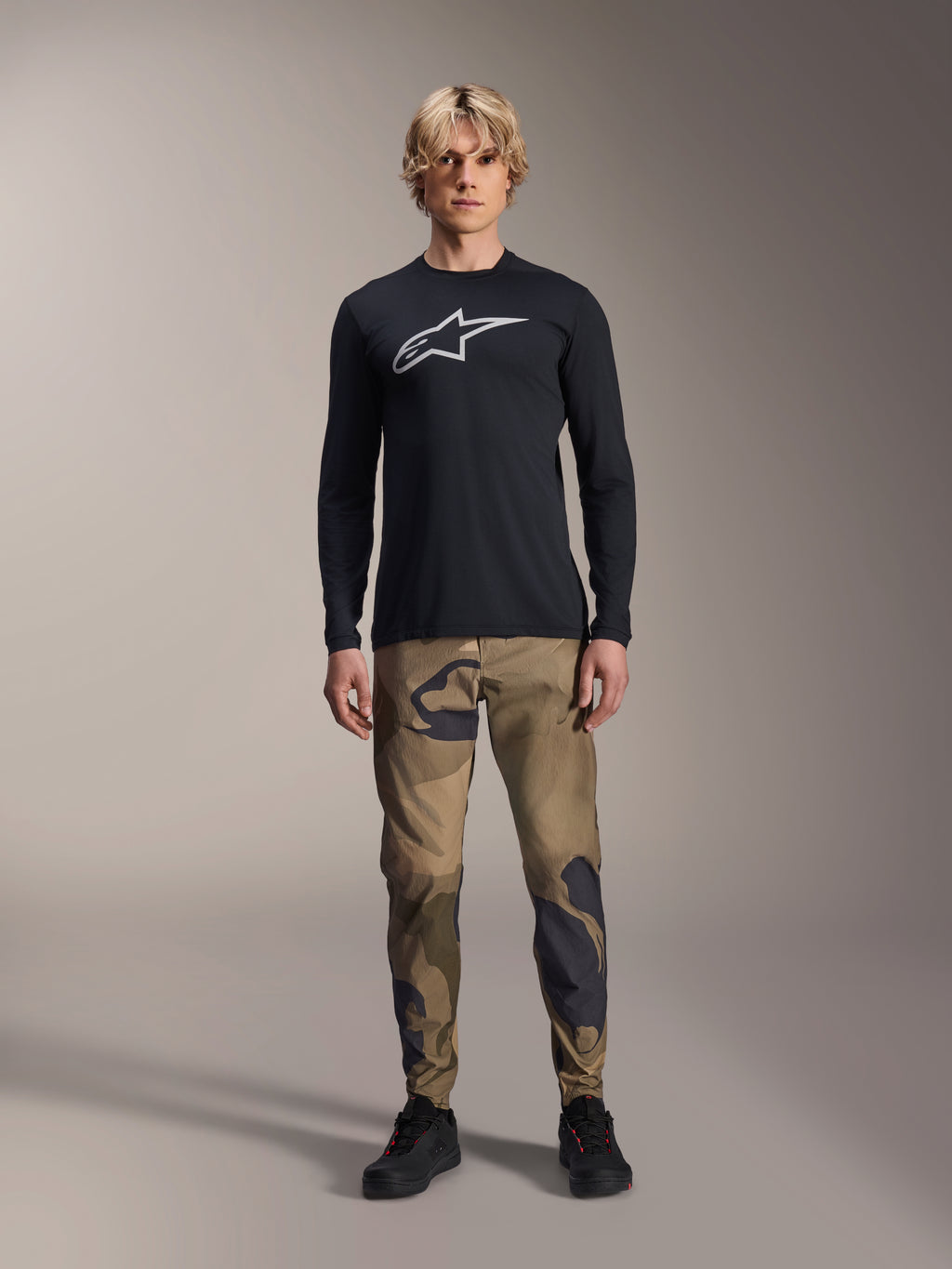Alpinestars A-Dura Astar Langarmtrikot, Schwarz, mit einem großen weißen Astars-Logo auf der Brust, getragen mit einer Camouflage-Hose und schwarzen Schuhen, lässige Mountainbike-Performance-Bekleidung