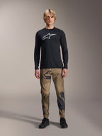 Alpinestars A-Dura Astar Langarmtrikot, Schwarz, mit einem großen weißen Astars-Logo auf der Brust, getragen mit einer Camouflage-Hose und schwarzen Schuhen, lässige Mountainbike-Performance-Bekleidung