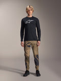 Alpinestars A-Dura Camo Hose, MTB-Radhose, braun-grünes Tarnmuster, kombiniert mit einem schwarzen Langarm-Jersey und schwarzen Schuhen, sportlicher Mountainbike-Stil aus strapazierfähigem Stretch-Material