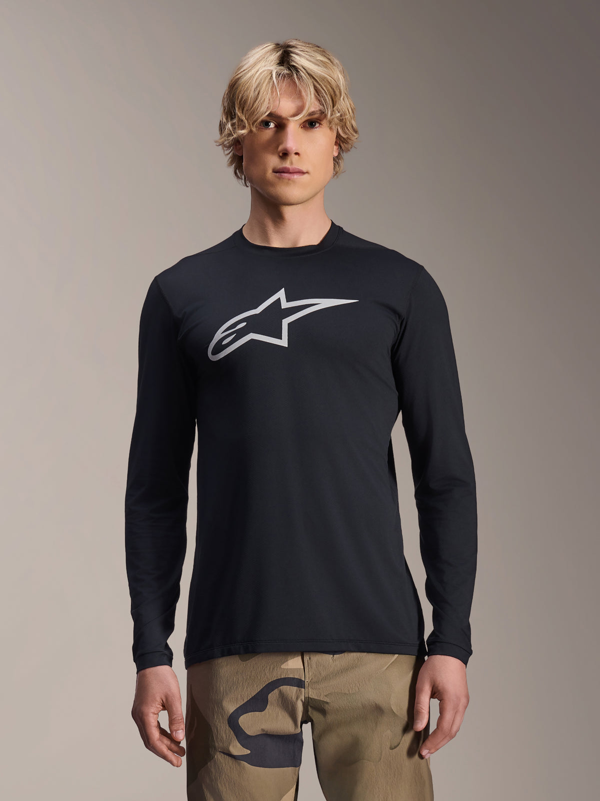 Alpinestars A-Dura Astar Langarm-Jersey, Schwarz mit silbernem Astars-Logo auf der Brust, getragen von einem Modell mit Camouflage-Hose, lässige MTB-Performance-Bekleidung für Mountainbiking