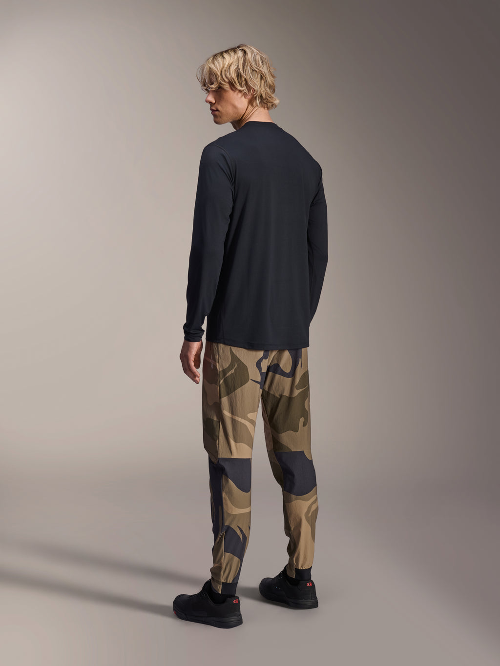 Alpinestars A-Dura Camo Hose, MTB-Hose, braunes Camouflage, getragen mit einem schwarzen Langarm-Jersey und schwarzen Sneakern, Rückansicht mit schmal zulaufenden Beinen und elastischen Bündchen, Performance-Bekleidung für den Offroad-Radsport
